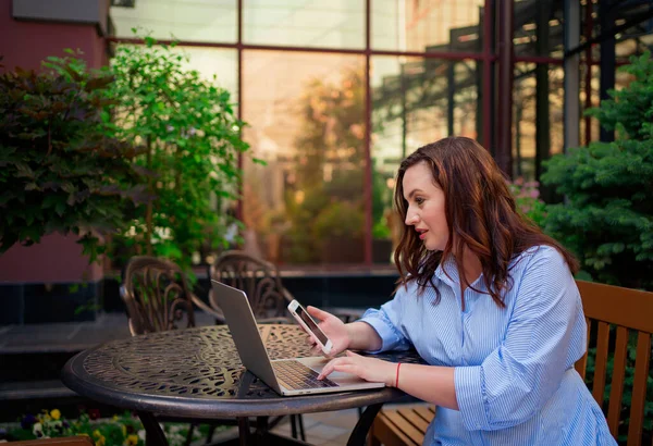 La mujer se sienta en una terraza al aire libre y trabaja en un ordenador portátil y un teléfono inteligente. — Foto de Stock