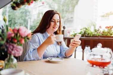 Mavi elbiseli çekici bir kadın kafeterya çayı içer ve akıllı telefondan mesaj atar.