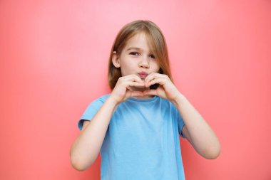 Pembe arka planda mavili küçük kız. Elleri kalp şeklinde katlanmış.