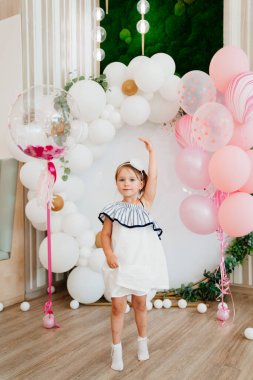 Beyaz elbiseli küçük bir kız doğum günü için balonlarla fotoğraf bölgesinde duruyor.