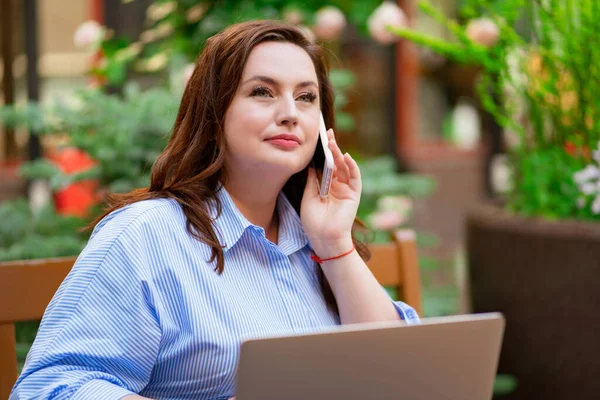 Kobieta siedzi na tarasie, pracuje na laptopie i rozmawia przez telefon — Zdjęcie stockowe