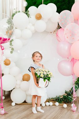 Bir buket çiçekli küçük bir kız elinde balonlarla fotoğraf bölgesinde duruyor.