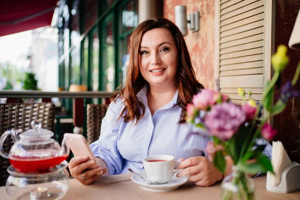 An attractive woman in a blue dress drinks cafe tea and texts in a smartphone — Stock Photo, Image