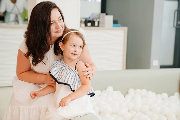 Madre e hija se sientan junto a la piscina seca con bolas blancas. sala de juegos para niños — Foto de Stock