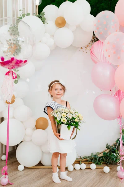 A little girlwith a bouquet of flowers stands on a photo zone with balloons