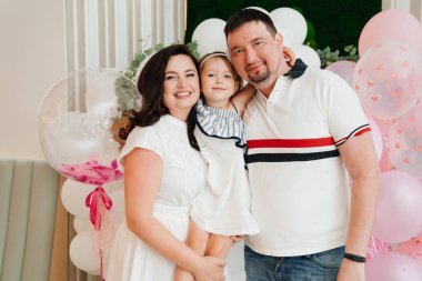 Annem, babam ve beyaz elbiseli küçük kız balonlarla fotoğraf bölgesinde duruyorlar.