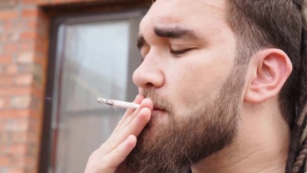 Крупным планом. Человек с бородой и дредами курит сигареты и выпускает дым — стоковое видео