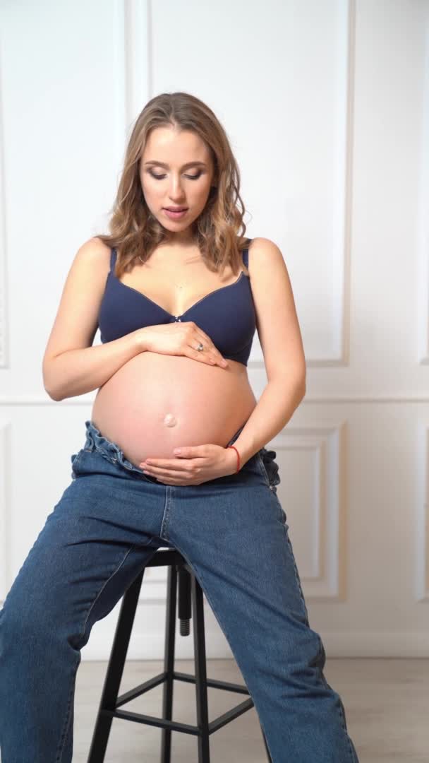 Verticale video. een aantrekkelijke zwangere vrouw met een kale buik zit op een stoel — Stockvideo