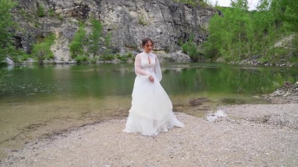 Hermosa novia en un vestido blanco se encuentra en la orilla de un lago de montaña. — Vídeos de Stock