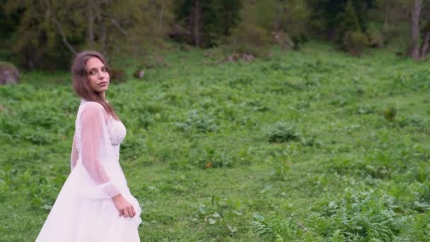 A young beautiful woman in a white wedding dress stands in a field with horses. — Stock Video