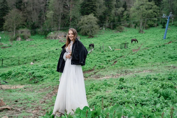 Mujer en un vestido de novia blanco y una chaqueta negra se encuentra en un campo con caballos. — Foto de Stock