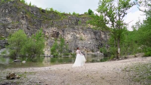 Novia en un vestido blanco con un ramo camina a la orilla de un lago de montaña. — Vídeo de stock