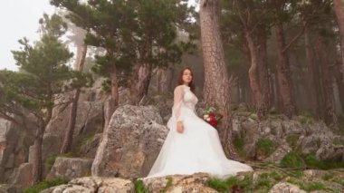 Gelin elinde bir buket kırmızı çiçekle dağ ormanında bir kayanın üzerinde duruyor..