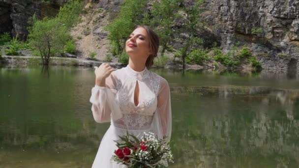 Krásná nevěsta v bílých šatech s kyticí na břehu horského jezera. — Stock video