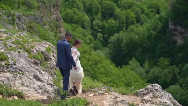 Braut und Bräutigam gehen in Hochzeitskleidern Händchen haltend in den Bergen. — Stockvideo