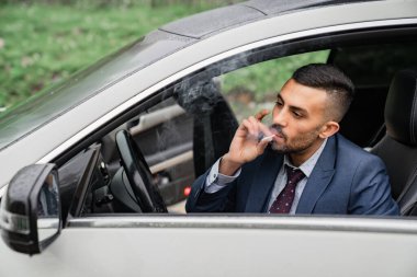 Bir adam arabanın direksiyonunda oturuyor, kapısı açık ve sigara içiyor..