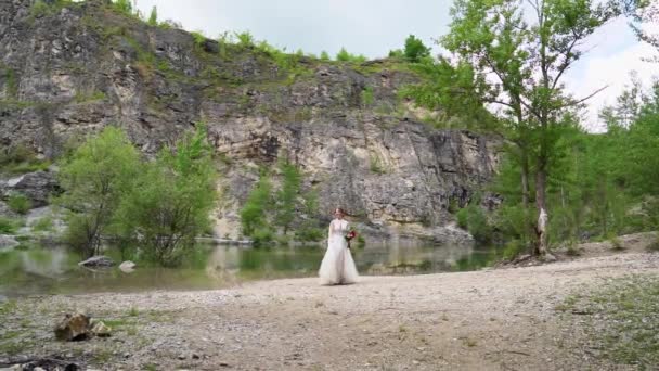 Bruid in een witte jurk met een boeket wandelingen op de oever van een bergmeer. — Stockvideo