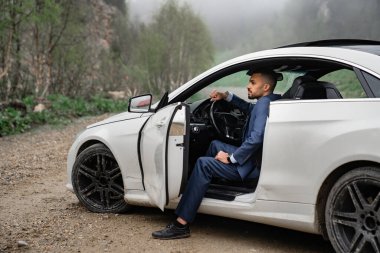Takım elbiseli çekici bir adam arabanın direksiyonunda oturuyor ve kapısı açık..
