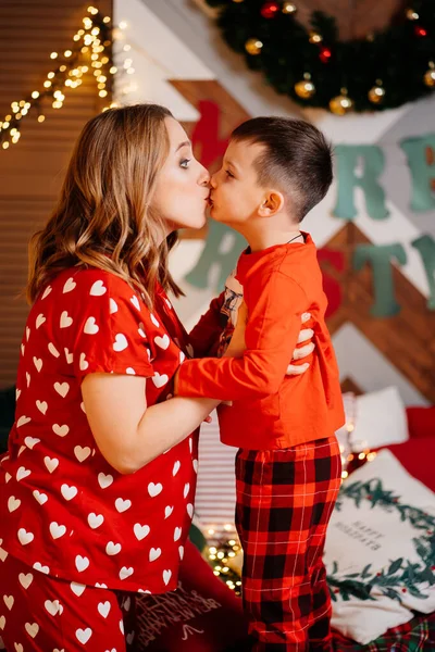 pregnant mother and son in red pajamas have fun on the bed. kiss