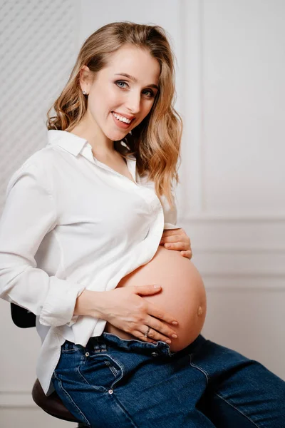 Uma mulher grávida atraente em uma camisa branca com uma barriga nua senta-se em uma cadeira. — Fotografia de Stock