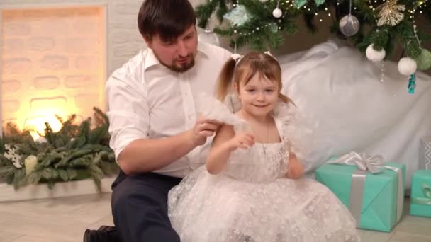 Ο μπαμπάς βοηθάει την κορούλα της να φορέσει ένα κομψό λευκό φόρεμα κάτω από το χριστουγεννιάτικο δέντρο. — Αρχείο Βίντεο