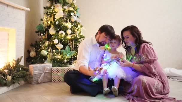 Apa, anya és kislánya ülnek a karácsonyfa mellett, és egy koszorúval játszanak.. — Stock videók