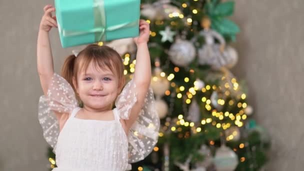 Vrolijk meisje in een wit jurk speelt met een geschenk doos in handen bij kerstboom — Stockvideo