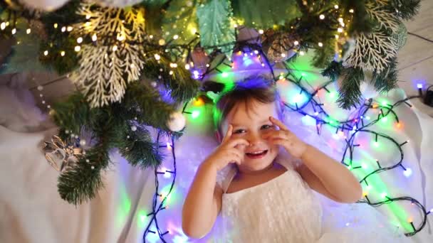 Ein süßes Mädchen liegt unter dem Weihnachtsbaum und spielt mit einem Kranz und Spielzeug — Stockvideo