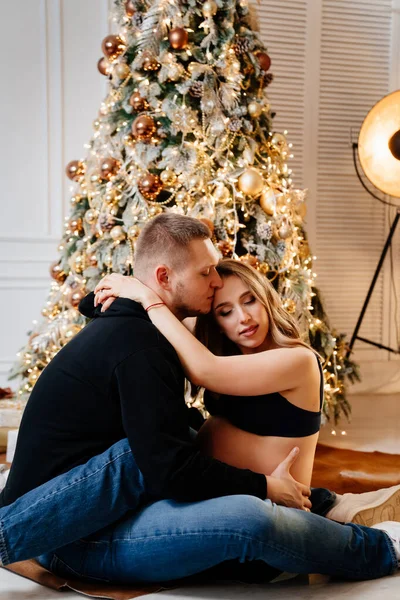 Homem feliz e mulher grávida sentado no chão pela árvore de Natal. — Fotografia de Stock