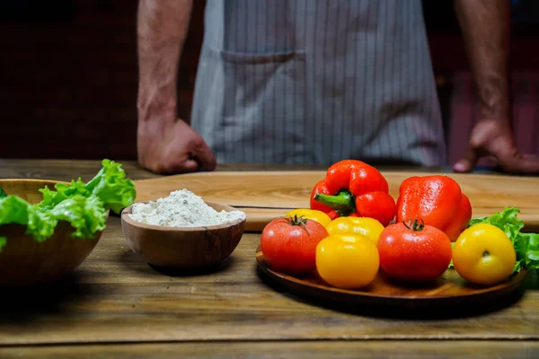 Uma tigela de madeira de farinha e legumes na mesa da cozinha. homem em um avental. — Fotografia de Stock