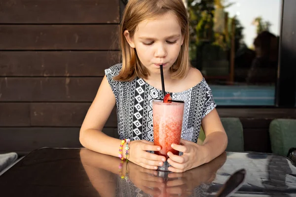 Funny little girl drinks through cocktail tube strawberry cocktail. family cafe, summer veranda