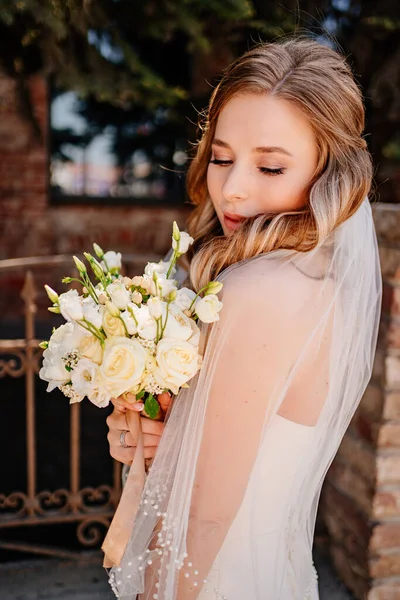 Sanfte und schöne Braut im Schleier mit einem Blumenstrauß am Zaun. — Stockfoto