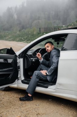 Bir adam arabanın direksiyonunda oturuyor, kapısı açık ve sigara içiyor..