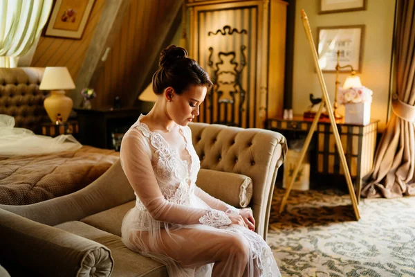 De ochtend van de bruid. mooie vrouw in kant badjas zitten op de bank in kamer. — Stockfoto