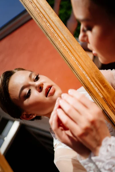 Uma bela jovem mulher em um manto de renda olha para seu reflexo no espelho. — Fotografia de Stock