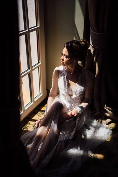 Een mooie en zachte vrouw in een sexy kant badjas zit bij het raam. — Stockfoto