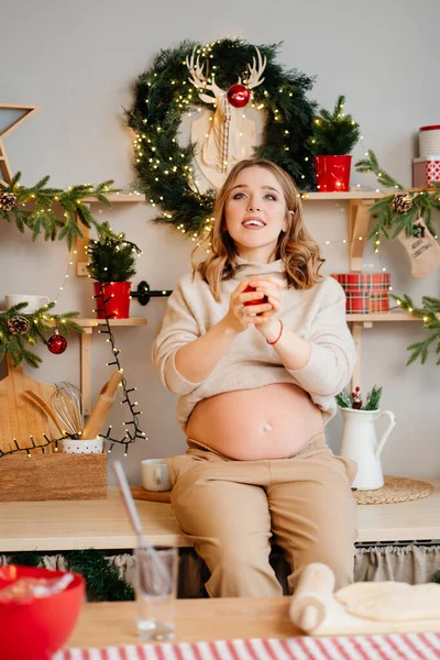 Μια όμορφη έγκυος γυναίκα κάθεται στο τραπέζι και ταχυδακτυλουργικά στην κουζίνα της Πρωτοχρονιάς. — Φωτογραφία Αρχείου