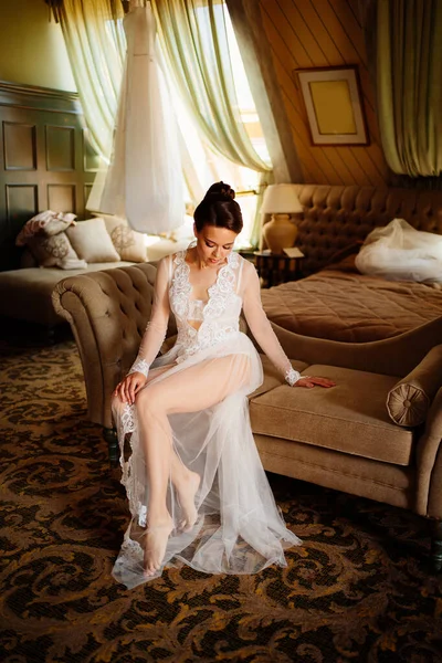 Mañana de la novia. hermosa mujer en túnica de encaje sentado en el sofá en la habitación. — Foto de Stock