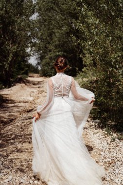 Gelinlikli gelin ağaçlar arasında dağ yolunda yürüyor. Arkadan görünüm. 