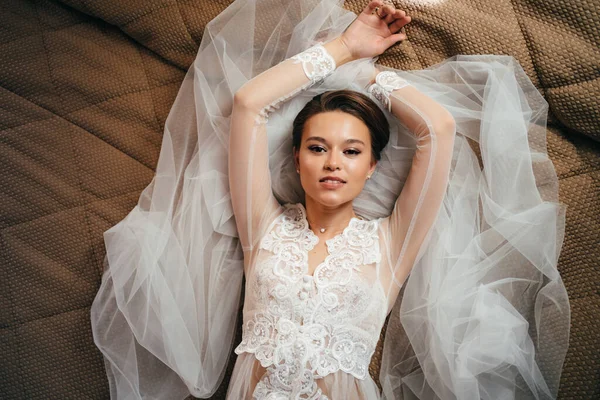 Hermosa novia en una túnica de encaje se encuentra en la cama sobre un velo. mañana antes de la boda — Foto de Stock