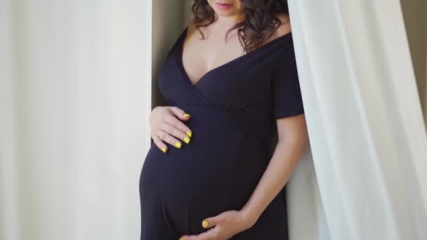 En gravid kvinna i svart klänning står vid fönstret med vita gardiner. — Stockvideo
