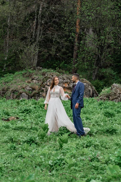 La mariée et le marié en vêtements de mariage marchent à travers la clairière en tenant la main. — Photo