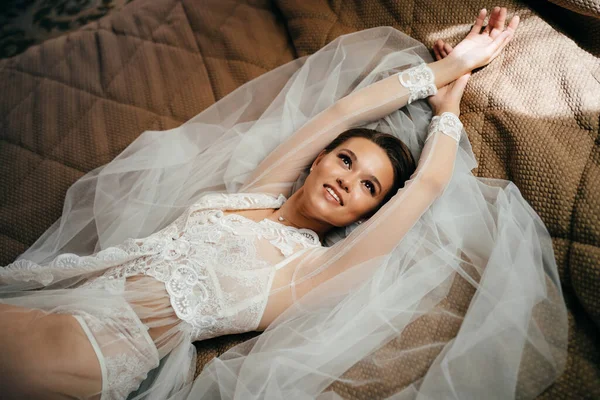 Hermosa novia en una túnica de encaje se encuentra en la cama sobre un velo. mañana antes de la boda — Foto de Stock