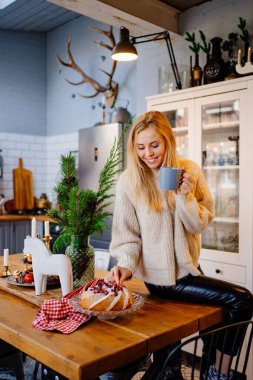 Yeni yıl süslemeleri için mutfakta çay içen güzel sarışın bir kadın..