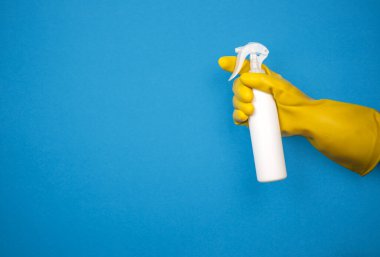 Sarı eldivenli bir el mavi arka planda bir dezenfektan spreyi tutar.