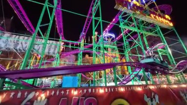 BUDAPEST, HUNGARY NOVEMBER 28, 2025: bir eğlence parkında gece çalışan neon ışıkları ile aydınlatılmış yüksek hızlı bir hız treni. Görüntüler parıldayan rayları yakalıyor ve karanlık akşam gökyüzüne karşı arabaları hareket ettiriyor..