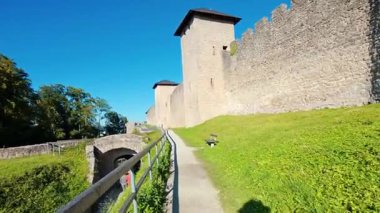 SALZBURG, AUSTRIA ECTOBER 03, 2025: Mavi bir gökyüzünün altındaki çimenli bir tepenin üzerinde bir gözetleme kulesi olan tarihi taş takviye duvarı duruyor. İnsanlar ortaçağ siperlerinin yakınındaki asfalt bir yolda yürüyor. Uzaktaki manzaralı vadi ve dağ manzaralı..