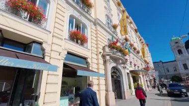 SALZBURG, AUSTRIA ECTOBER 3, 2025: Otel Bristol 'un tarihi cephesinde kırmızı çiçekler ve sarı bayraklarla süslü balkonlar yer almaktadır. Yayalar güneşli bir günde klasik Avrupa mimarisinin önünde kaldırım boyunca yürürler..