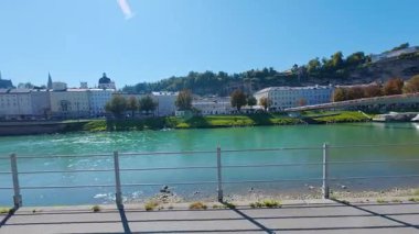 SALZBURG, AUSTRIA ECTOBER 03, 2025: Arkaplanda tarihi binalar ve tepeler bulunan Salzach Nehri 'nin geniş bir görüntüsü. Sahnede bir nehir kenarı geçidi ve açık mavi gökyüzünün altında bir yaya köprüsü yer alıyor..