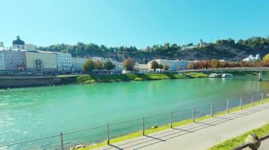 SALZBURG, AUSTRIA ECTOBER 03, 2025: Arkaplanda tarihi binalar ve tepeler bulunan Salzach Nehri 'nin geniş bir görüntüsü. Sahnede bir nehir kenarı geçidi ve açık mavi gökyüzünün altında bir yaya köprüsü yer alıyor..